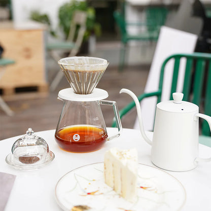 Coffee brewing setup with a pour-over coffee maker, white teapot, and slice of cake on a table.