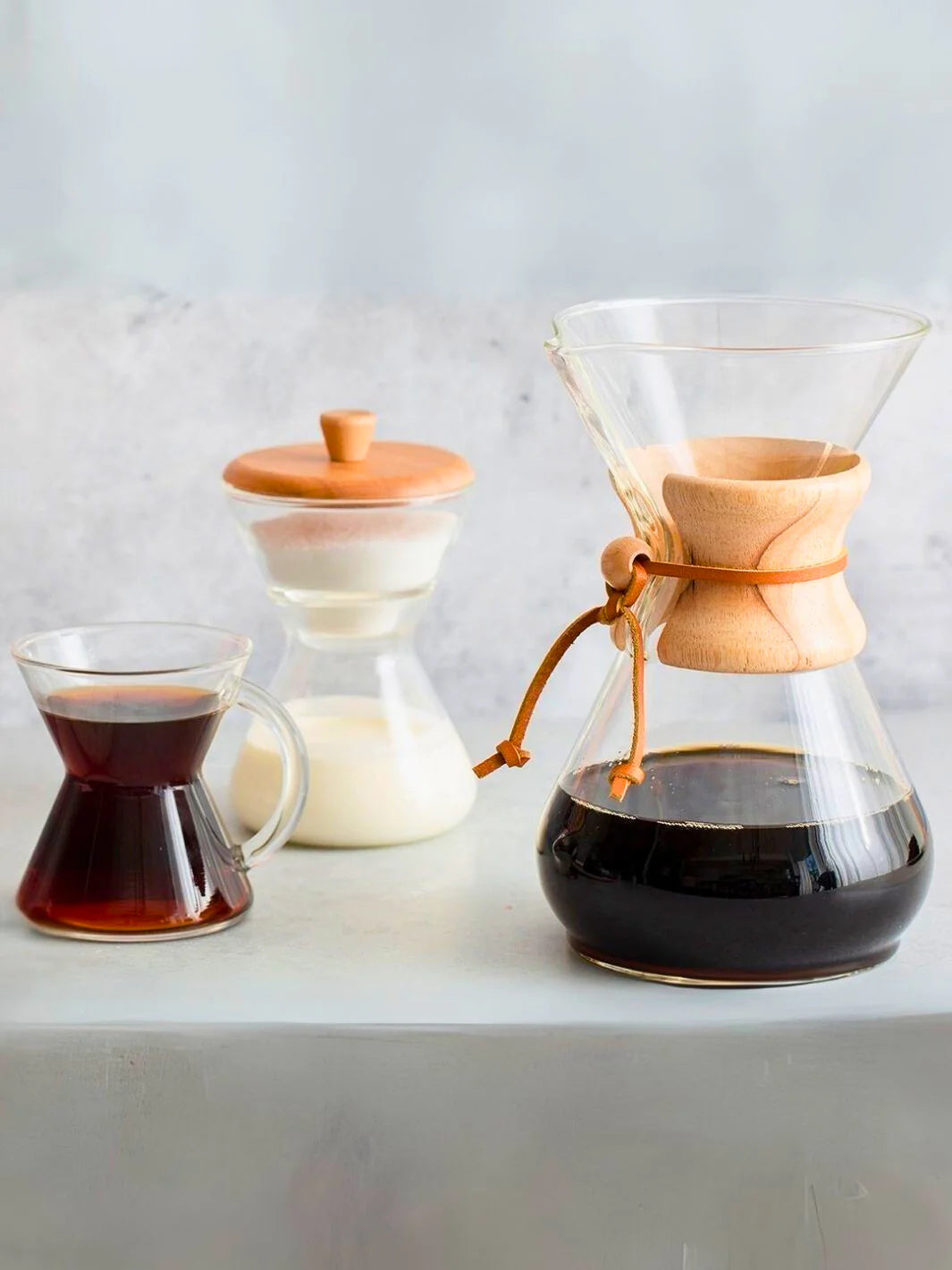 Set of coffee-making equipment including a glass carafe, wooden lid, and a small glass mug with coffee on a light background.