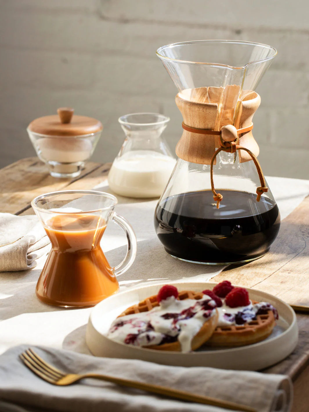 Coffee-making setup with a Chemex coffee maker, a cup of coffee, and a plate of waffles with berries on a wooden table.