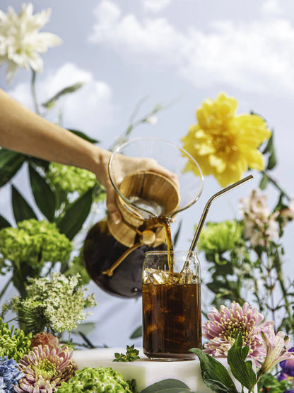 Person pouring a drink into a glass with a straw, surrounded by flowers outdoors.