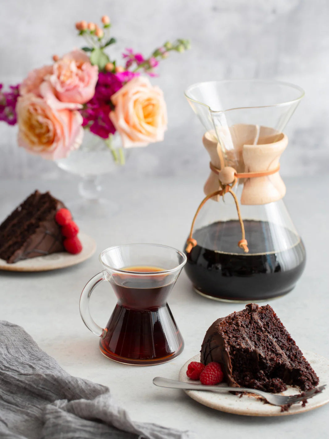 Chemex coffee maker with a cup of coffee, chocolate cake, and flowers on a light surface.