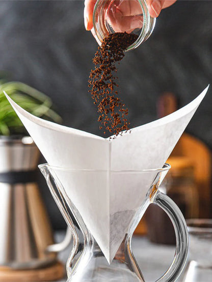 Person pouring coffee grounds into a white filter paper over a glass coffee maker.