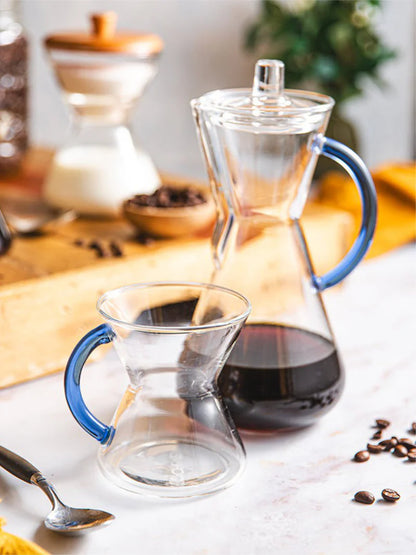 Clear glass coffee carafe with blue handle and glass mug on a wooden surface with coffee beans and a spoon.