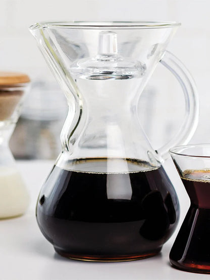 Clear glass carafe filled with coffee next to a small glass of coffee on a light background