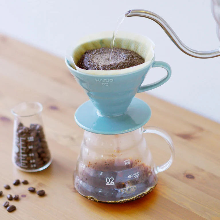 Coffee brewing process using a turquoise Hario coffee dripper on a wooden surface with coffee beans and a glass container.