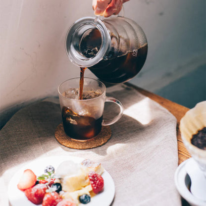 Person pouring coffee from a carafe into a glass mug on a table with a plate of fruit.