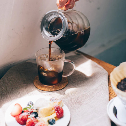 Person pouring coffee from a carafe into a glass mug on a table with a plate of fruit.