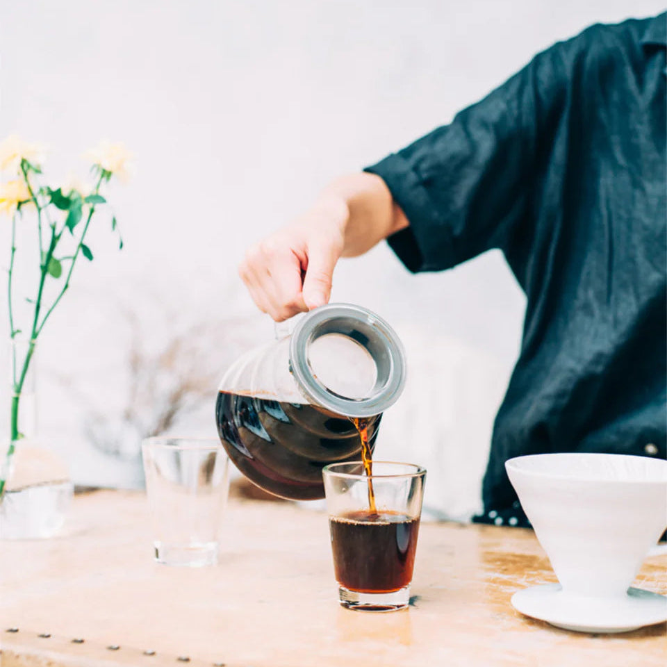 Person pouring tea from a glass teapot into a small glass on a wooden table.